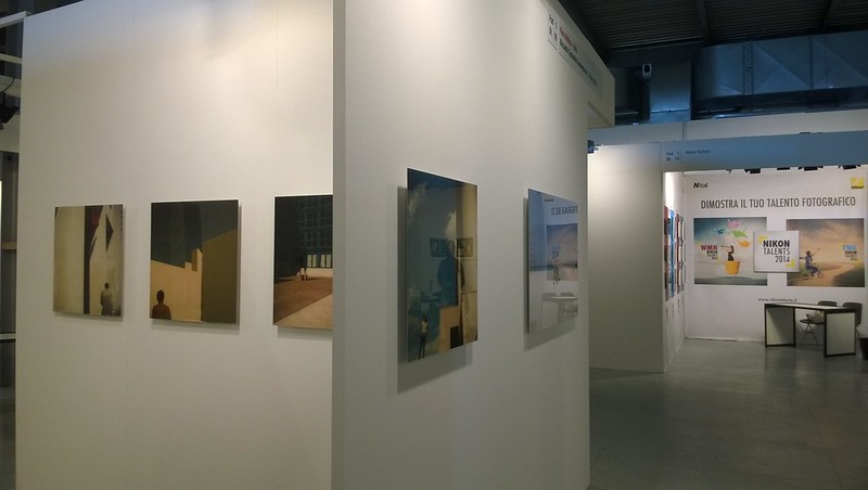 View down a white-walled corridor at an art exhibition, featuring minimalist paintings with figures and architectural elements. A sign for "NIKON TALENTS 2014" is visible in the background.