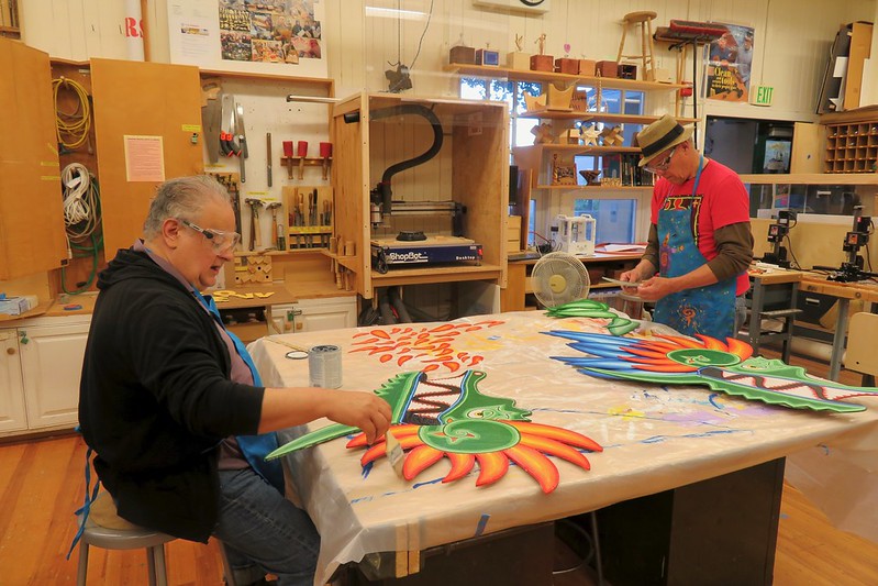 Two artists are working in a cluttered studio space. One seated artist is painting a colorful wooden cutout, while another standing artist is working at a nearby table. Tools, supplies, and finished pieces are visible throughout the workshop.