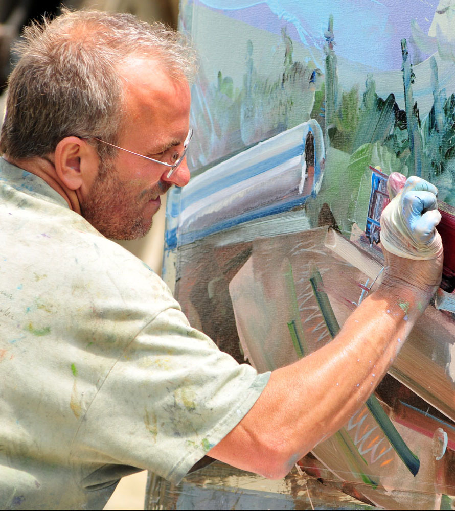 Close-up of David Brewster, wearing glasses and a paint-splattered shirt, intensely focused on painting on a canvas outdoors. He is using a palette knife with blue paint.