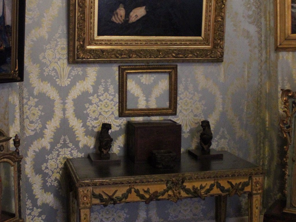Ornate table against a patterned wallpaper with two dark sculptures flanking a small box, and framed artwork above.