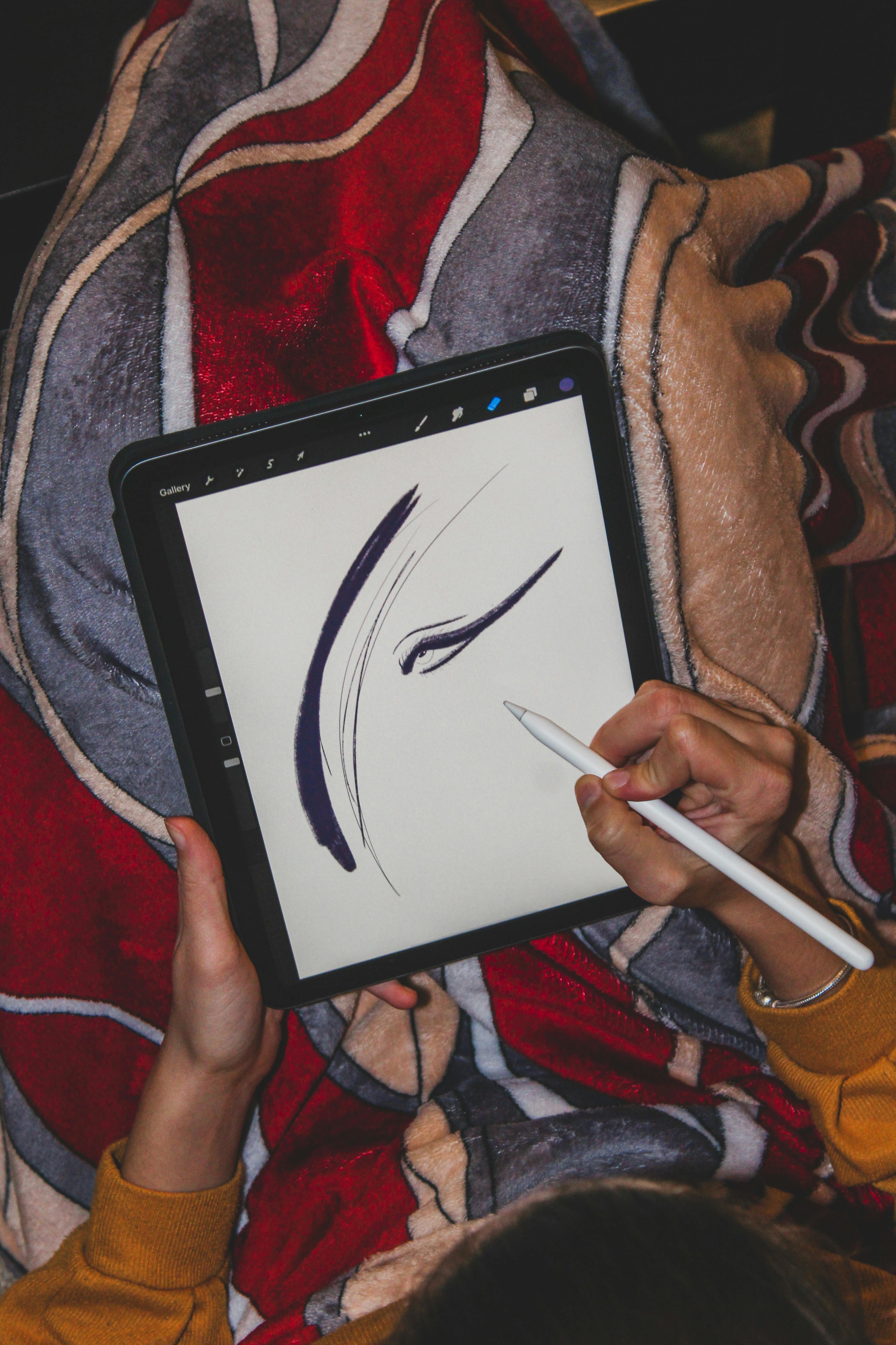 A person's hands holding a stylus and drawing on a digital tablet, with a blanket in the background.